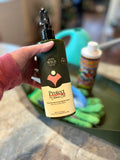 A hand holding a bottle of Protect Neem Oil against a blurred background with a teal watering can and other houseplant accessories at Paisley Gardens in Northfield, MN.