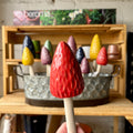 A hand holds a glossy red morel mushroom garden stake with a textured surface and a light wood stick. Behind it, a wooden crate and galvanized metal tub hold several morel mushroom in different colors, including yellow, blue, green, purple, and red against a rustic indoor backdrop. At Paisley Gardens in Northfield, MN.