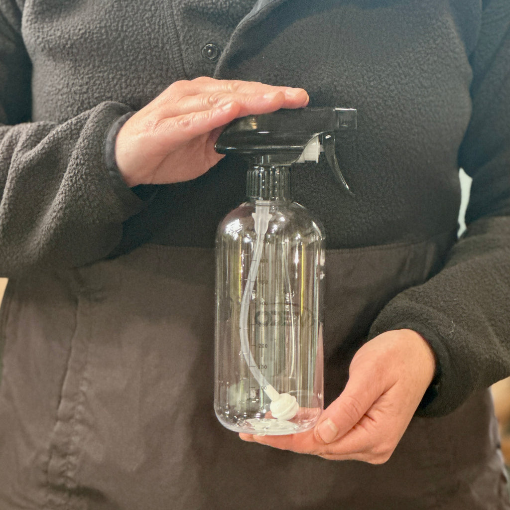 A person wearing a dark brown fleece jacket holds a clear plastic spray bottle with a black trigger sprayer and a white internal tube at Paisley Gardens in Northfield, MN.
