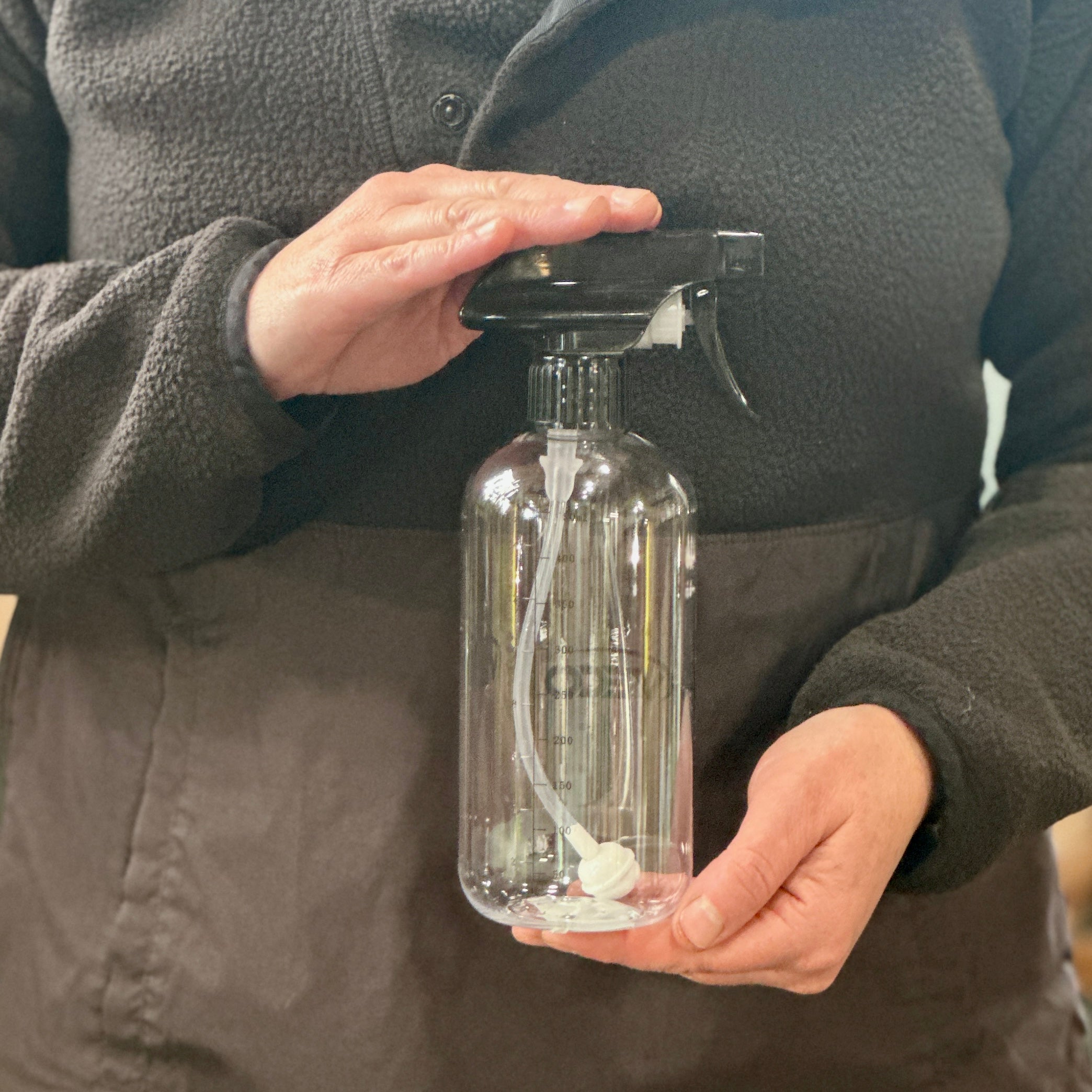 A person wearing a dark brown fleece jacket holds a clear plastic spray bottle with a black trigger sprayer and a white internal tube at Paisley Gardens in Northfield, MN.