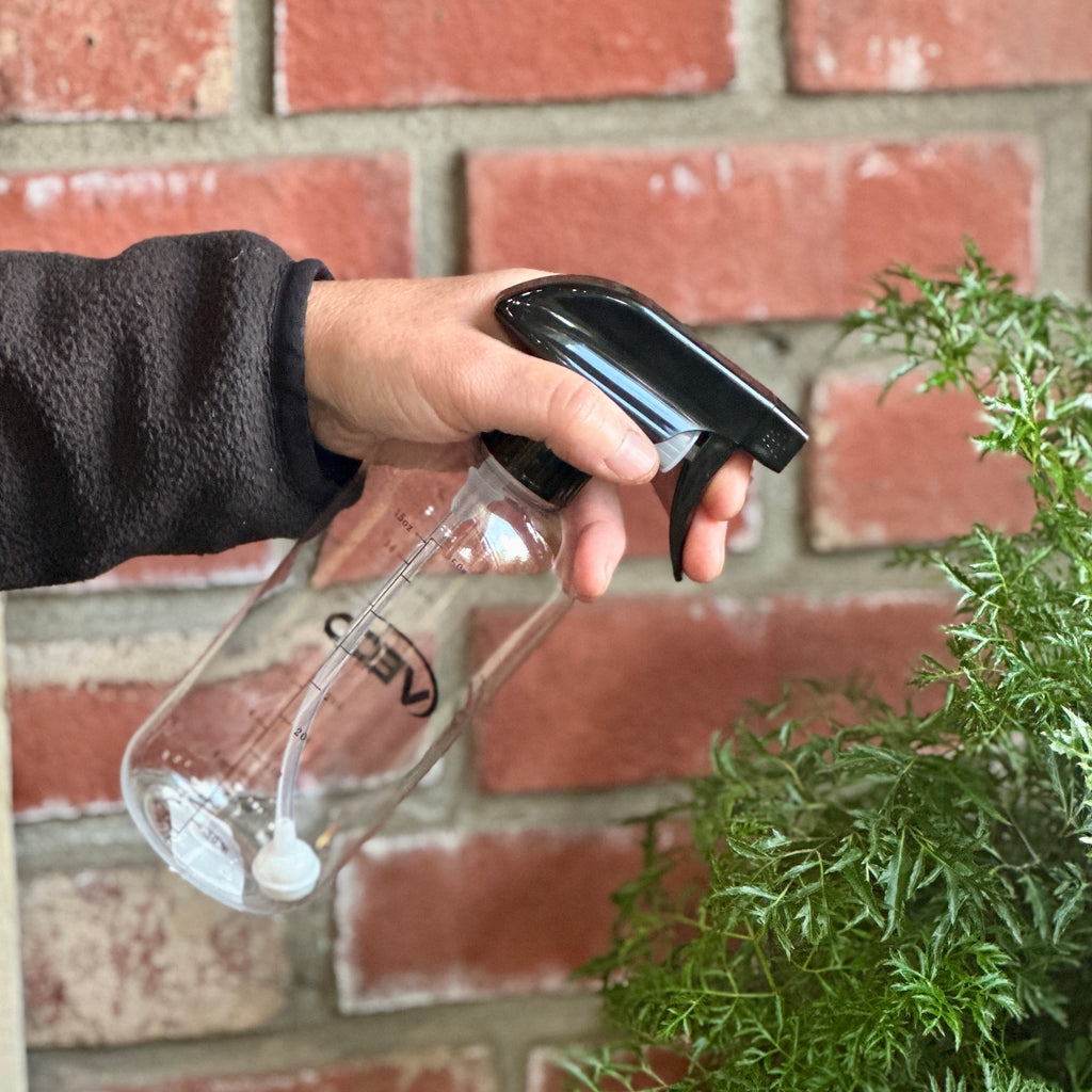 A hand holding a clear plastic spray bottle with a black trigger sprayer, spraying water onto a green plant in front of a red brick wall at Paisley Gardens in Northfield, MN