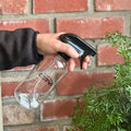 A hand holding a clear plastic spray bottle with a black trigger sprayer, spraying water onto a green plant in front of a red brick wall at Paisley Gardens in Northfield, MN