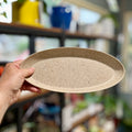 A hand holding a speckled natural colored, oval tray in a brightly lit, blurred plant room at Paisley Gardens in Northfield, MN.