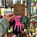 A hand holding a pair of pink, white and black gardening gloves in front of shelves filled with plants and pots at Paisley Gardens in Northfield, MN.
