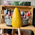 A yellow textured morel mushroom garden stake with a wooden handle, displayed in front of a wooden crate holding other colorful morel mushroom garden stakes. at Paisley Gardens in Northfield, MN.