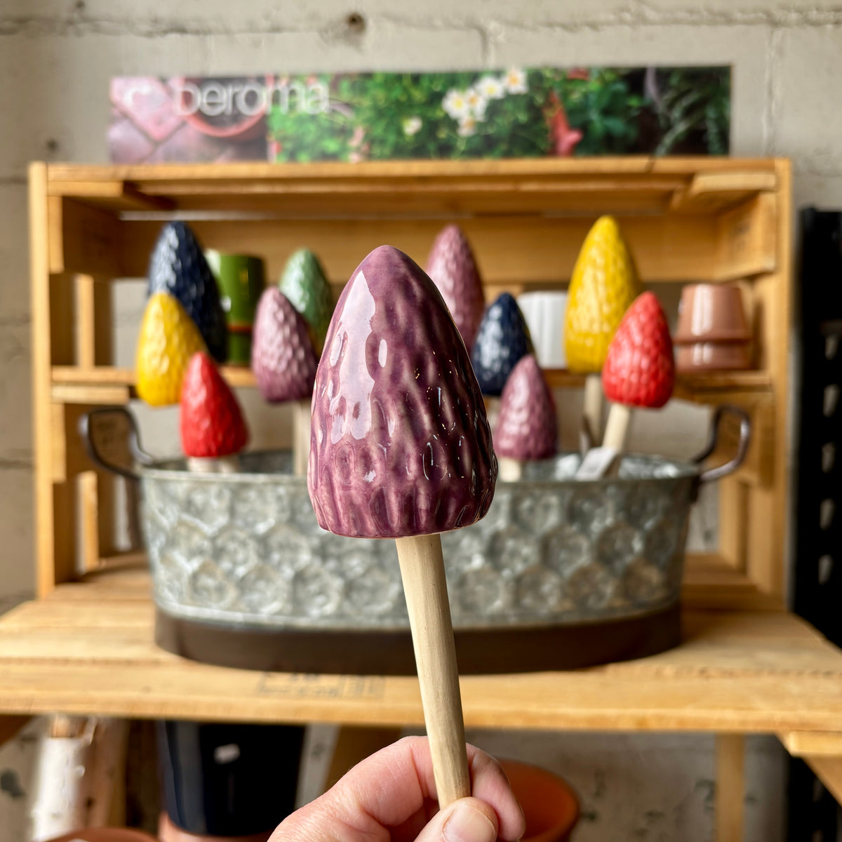 A hand holds a glossy purple morel mushroom garden stake with a wooden stick. A metal tub of colorful mushroom stakes sitting on a wooden shelf in the background at Paisley Gardens in Northfield, MN.