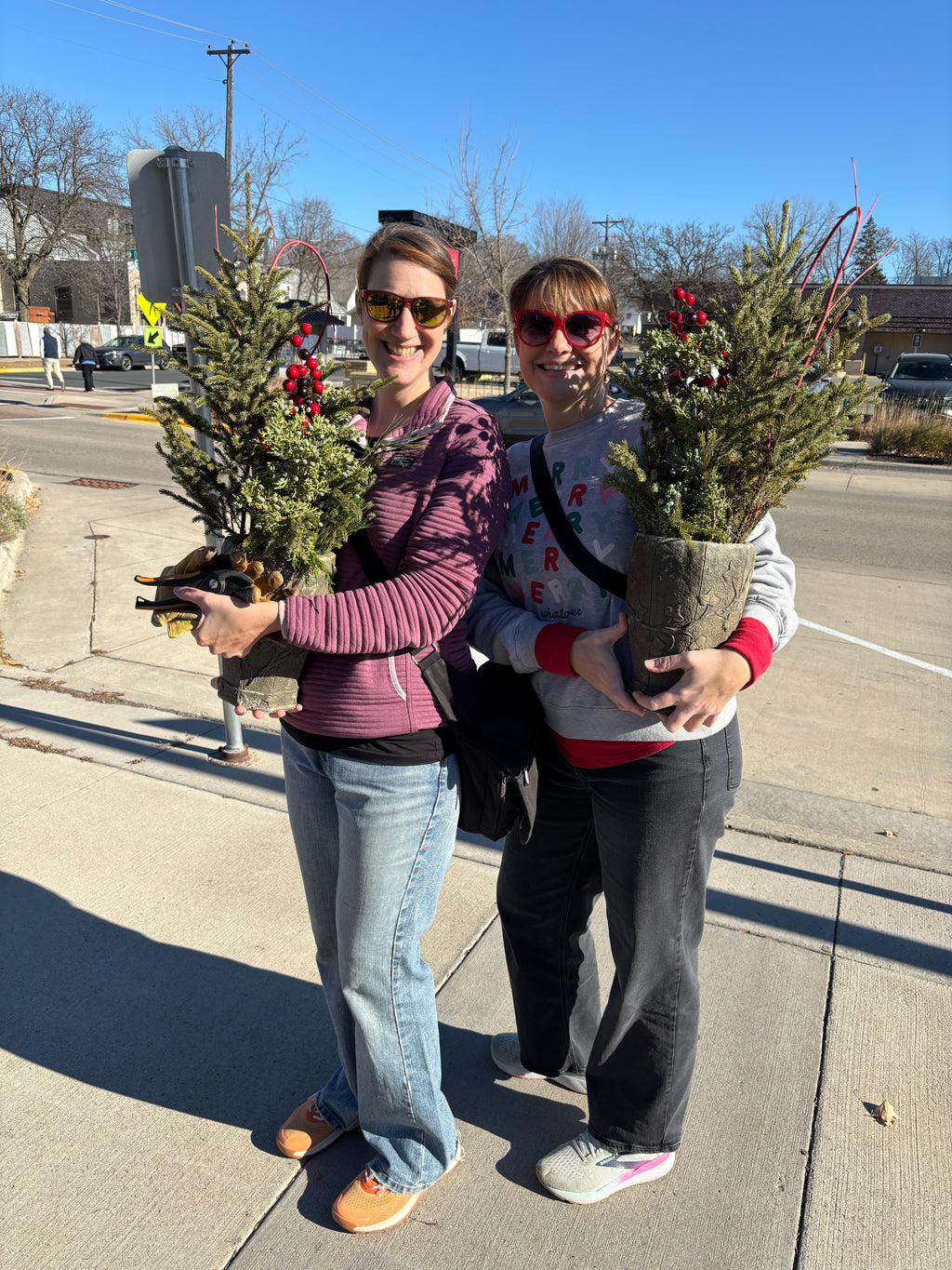 Two women standing on a sunny sidewalk outdoors, each holding a small potted Christmas pot with green branches and red berry decorations. One wears a purple striped zip-up top and blue jeans, and the other wears a gray graphic sweatshirt and dark pants at Paisley Gardens in Northfield, MN.